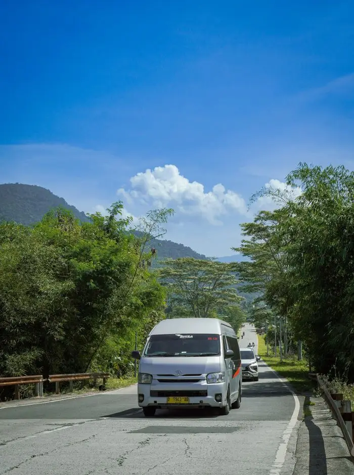 our-experience White minivan driving down a lush, scenic road in Garut, West Java, Indonesia under a vibrant blue sky.